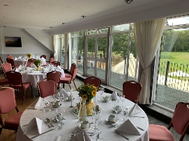 Tables set out for a lunch with white tablecloths, flowers, cutlery etc. In the background, there is the view of the golf course.